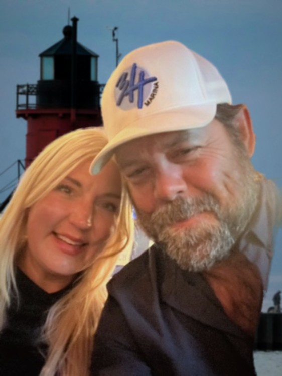Katelyn Hubbard and Jeffrey R. Hubbard in front of a red Great Lakes pier lighthouse at dusk, Jeffrey wearing the 3H Marina cap.